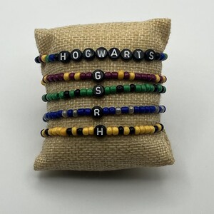 Könnte beinhalten: Sechs Armbänder mit Perlen, auf denen schwarze Buchstabenperlen "HOGWARTS", "G", "S", "R" und "H" stehen. Die Armbänder sind in verschiedenen Farben gehalten, darunter Gold, Grün, Blau und Burgunderrot, und werden auf einem Jutekissen präsentiert.