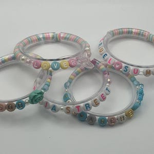May include: Five clear acrylic bracelets with colourful beads spelling out words like "LOVER", "GOOD", "SUMMER", and "ARCHIE". The bracelets have pastel-coloured accents and decorative charms, perfect for a fun, personalised accessory.