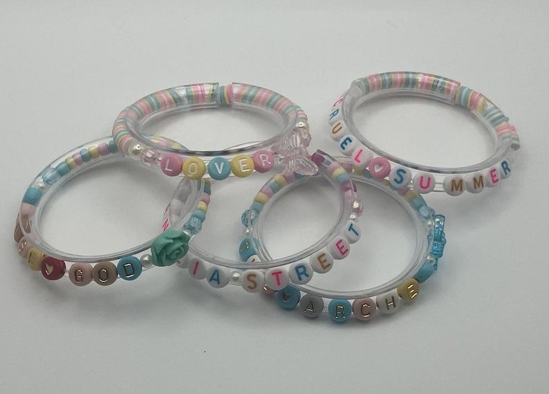 May include: Five clear acrylic bracelets with colourful beads spelling out words like "LOVER", "GOOD", "SUMMER", and "ARCHIE". The bracelets have pastel-coloured accents and decorative charms, perfect for a fun, personalised accessory.