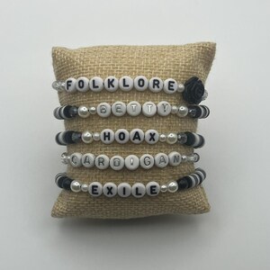 K&ouml;nnte beinhalten: Vier Armb&auml;nder mit Perlen mit wei&szlig;en Buchstaben, die "FOLKLORE", "BETTY", "HOAX", "CARDIGAN" und "EXILE" buchstabieren. Die Armb&auml;nder haben schwarze und wei&szlig;e Perlen und werden auf einem Jutekissen pr&auml;sentiert.