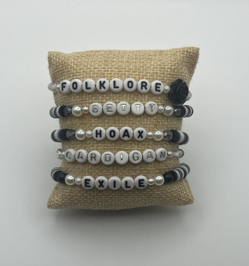 K&ouml;nnte beinhalten: Vier Armb&auml;nder mit Perlen mit wei&szlig;en Buchstaben, die "FOLKLORE", "BETTY", "HOAX", "CARDIGAN" und "EXILE" buchstabieren. Die Armb&auml;nder haben schwarze und wei&szlig;e Perlen und werden auf einem Jutekissen pr&auml;sentiert.