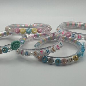 May include: Five clear plastic bangles with colourful beads spelling out words such as "LOVE", "WELCOME SUMMER", and "LIA STREET". Some bangles have decorative accents like a rose or a butterfly.