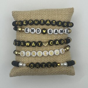 May include: Five beaded bracelets with black, white, and gold beads. The bracelets spell out the words "REPUTATION", "END GAME", "AT AWAY CAR", "GORGEOUS", and "DRESS". The bracelets are displayed on a burlap pillow.