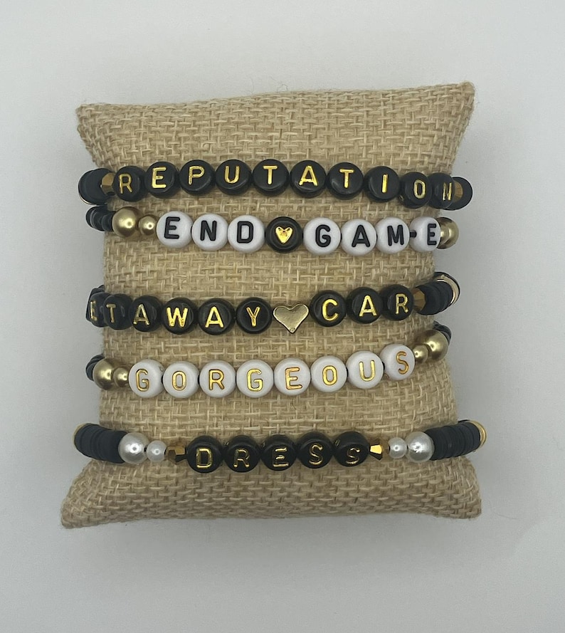 May include: Five beaded bracelets with black, white, and gold beads. The bracelets spell out the words "REPUTATION", "END GAME", "AT AWAY CAR", "GORGEOUS", and "DRESS". The bracelets are displayed on a burlap pillow.
