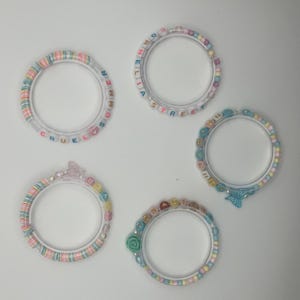 May include: Four clear acrylic bracelets with colourful beads. One bracelet spells "CRUEL SUMMER", another "CORNE LIA STREET", a third "EVER", and the last "SHE". Each bracelet has a unique design, including a butterfly and a rose.