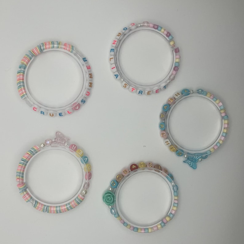 May include: Four clear acrylic bracelets with colourful beads. One bracelet spells "CRUEL SUMMER", another "CORNE LIA STREET", a third "EVER", and the last "SHE". Each bracelet has a unique design, including a butterfly and a rose.