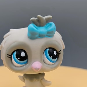 May include: A small gray and white toy bird with blue eyes and a blue bow on its head. The bird has pink feet and is standing on a yellow surface.