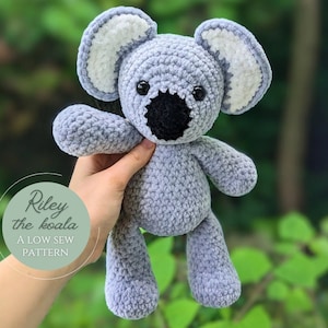 May include: A crocheted gray koala plush toy with white ears and black eyes. The toy is being held by a hand and the text "Riley the koala A LOW SEW PATTERN" is visible in the background.