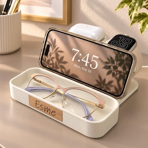 Op de afbeelding: Een wit geribbeld oplaadstation met een houten inlegstuk met de naam Esme. Het apparaat houdt een smartphone vast die 7:45 weergeeft, witte draadloze oordopjes en een smartwatch. Een bril met gouden montuur ligt in het voorste bakje.