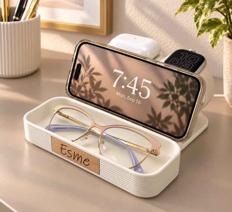 Op de afbeelding: Een wit geribbeld oplaadstation met een houten inlegstuk met de naam Esme. Het apparaat houdt een smartphone vast die 7:45 weergeeft, witte draadloze oordopjes en een smartwatch. Een bril met gouden montuur ligt in het voorste bakje.