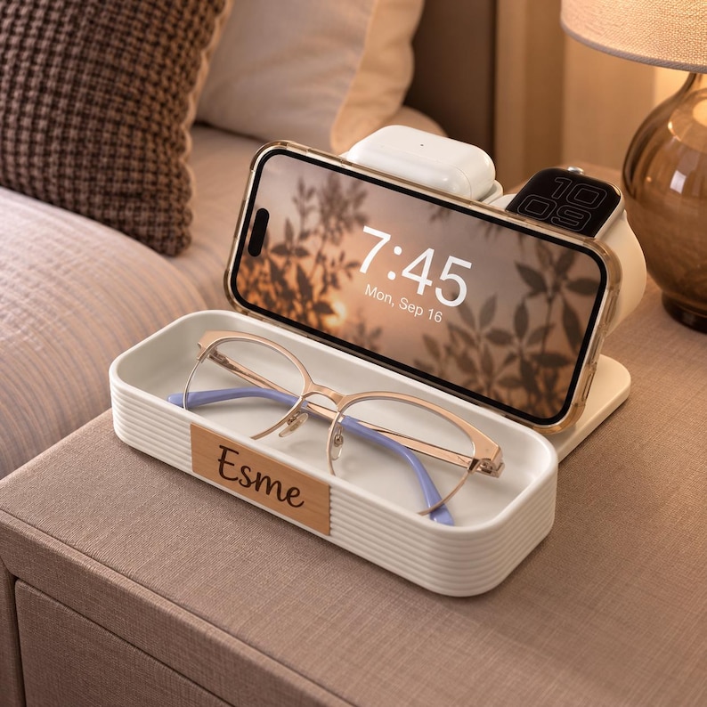 Op de afbeelding: Een wit oplaadstation voor meerdere apparaten met een geribbelde buitenkant en een houten accentplaat met de naam Esme. De standaard houdt een smartphone met 7:45 in beeld, draadloze oordopjes, een smartwatch en een bakje voor een bril.