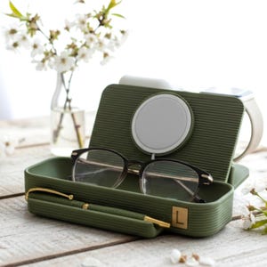 May include: An olive green glasses case with a matching pen and a pair of black-rimmed glasses. The case is open, revealing a white circular charging pad. A gold-colored letter "L" is on the case. A white smartwatch is in the background.