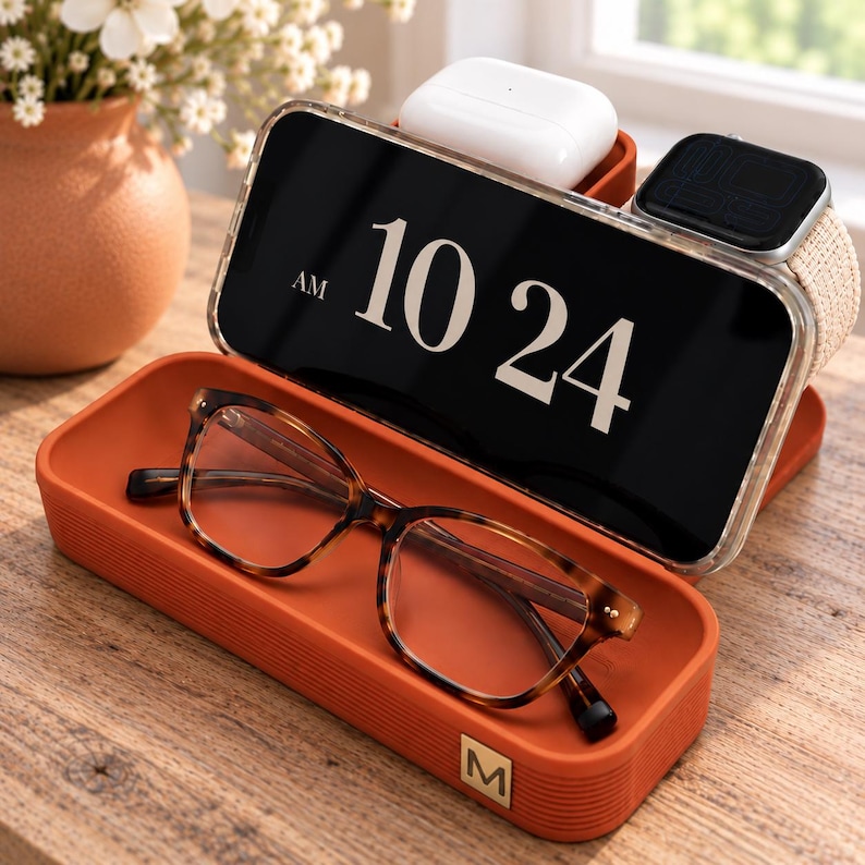 Op de afbeelding: Een rechthoekig bureau-organizerbakje in terracotta-kleur met een klein goudkleurig M-logo. Het bakje bevat een smartphone met de tijd 10:24 AM, een bril met schildpadpatroon, een witte oplaadcase voor oordopjes en een smartwatch.
