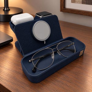 Op de afbeelding: Een marineblauw oplaadstation voor meerdere apparaten met een geribbelde textuur, voorzien van een ronde magnetische oplader, een opbergvak voor accessoires, een witte draadloze oordopjescase, een smartwatch en een zilverkleurige pilotenbril.
