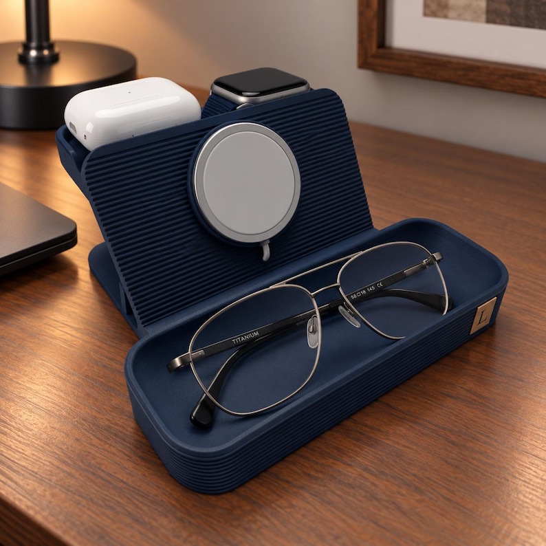 Op de afbeelding: Een marineblauw oplaadstation voor meerdere apparaten met een geribbelde textuur, voorzien van een ronde magnetische oplader, een opbergvak voor accessoires, een witte draadloze oordopjescase, een smartwatch en een zilverkleurige pilotenbril.