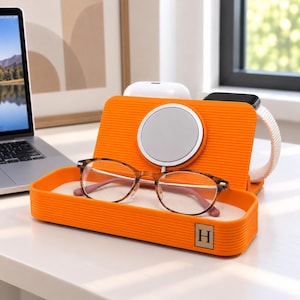 Op de afbeelding: Een oranje oplaadstation met een geribbelde textuur en een vierkant H-logo label. Het beschikt over een ronde magnetische oplader, een bakje voor accessoires en een gleuf voor een horloge. Een bril met schildpadpatroon rust in het bakje, met witte oordopjes en een horloge zichtbaar achter het oplaadpaneel.