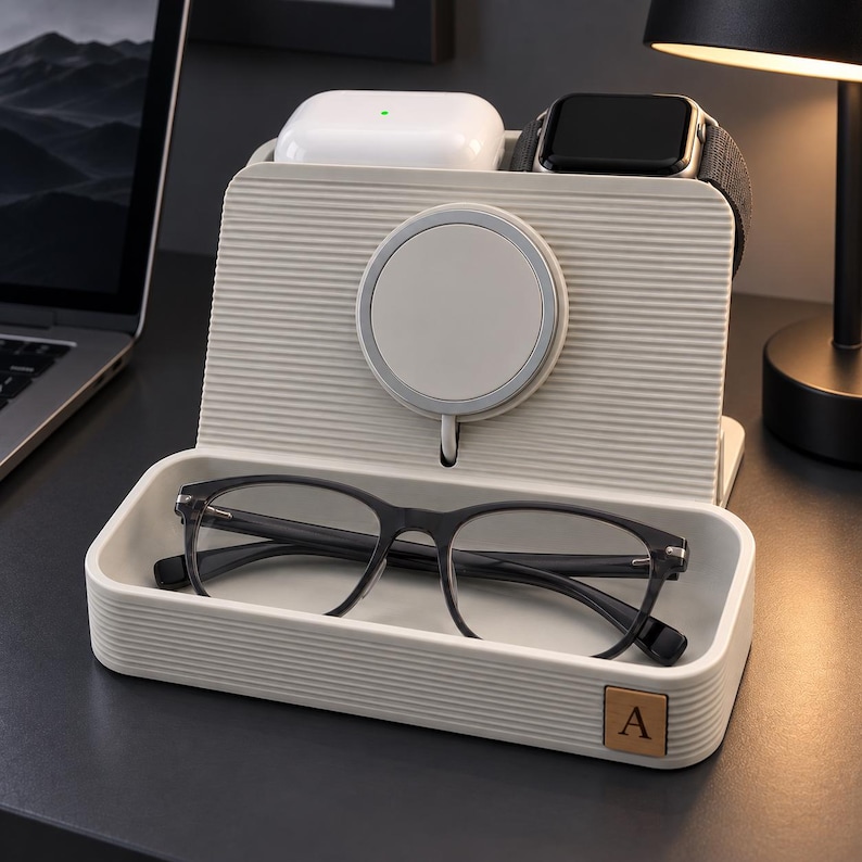 Op de afbeelding: Een beige, geribbeld oplaadstation met een ronde magnetische oplader, een bakje voor een bril en vakjes voor een witte oordopjescase en een zwarte smartwatch. Een klein vierkant label met de letter A is bevestigd aan de rechtervoorkant van de lade.