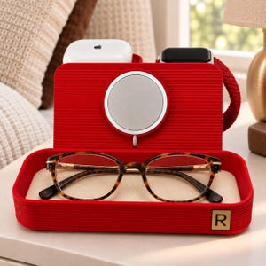 Op de afbeelding: Een rood oplaadstation voor meerdere apparaten met een geribbelde textuur en een vierkant R-logo. Het bevat een ronde magnetische oplader, een witte draadloze oordopjescase, een zwarte smartwatch en een bruine bril met schildpadpatroon.