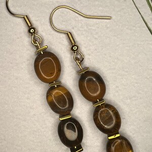May include: A pair of gold earrings with brown tiger's eye gemstone beads. Each earring has three beads and a gold hook.
