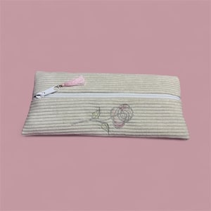 Può includere: Un astuccio per matite in velluto a coste beige con una cerniera bianca e un nappa rosa. L'astuccio presenta un ricamo a forma di rosa rosa e verde. L'astuccio è rettangolare e ha una consistenza morbida.