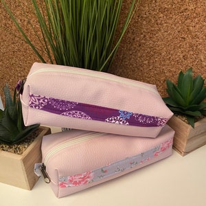 Può includere: Due pochette rosa con cerniera e bordi in tessuto con motivi diversi. La pochette superiore ha un motivo floreale viola e blu, e la pochette inferiore ha un motivo floreale rosa e grigio.