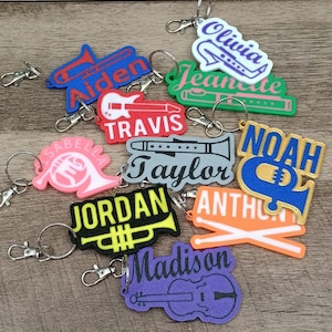 May include: A collection of colorful keychains featuring musical instruments and names. Keychains include a trumpet, guitar, flute, french horn, and violin. Names include Aiden, Travis, Olivia, and Madison. Each keychain has a silver clasp.