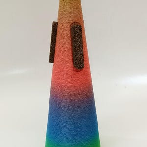 May include: A colorful cone-shaped vase with a textured surface. The vase is decorated with a gradient of yellow, orange, pink, blue, and green. It has two brown rectangular pieces attached to the side.