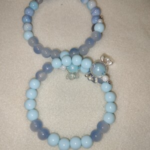 May include: Two beaded bracelets. One bracelet features light blue and gray beads, while the other has light blue beads and a candy-shaped charm. The bracelets are connected by a cluster of light blue beads and a small charm.
