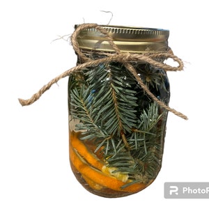 May include: A clear glass jar filled with evergreen sprigs and orange slices, tied with twine. The jar has a gold-coloured lid and is filled with a clear liquid. The twine is tied in a bow around the jar's neck. The jar is a decorative item.