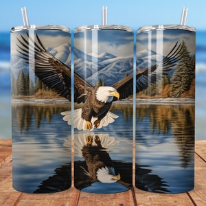 Peut inclure: Un gobelet en acier inoxydable avec un motif d'un aigle chauve volant au-dessus d'un lac avec des montagnes en arrière-plan. L'aigle se reflète dans l'eau.