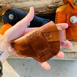Op de afbeelding: Een bruin leren holster met decoratieve stiksels en een riem. Andere holsters in zwart en oranje zijn zichtbaar op de achtergrond. De holsters zijn gemaakt van leer en hebben metalen knoopsluitingen. De stiksels zijn zichtbaar.