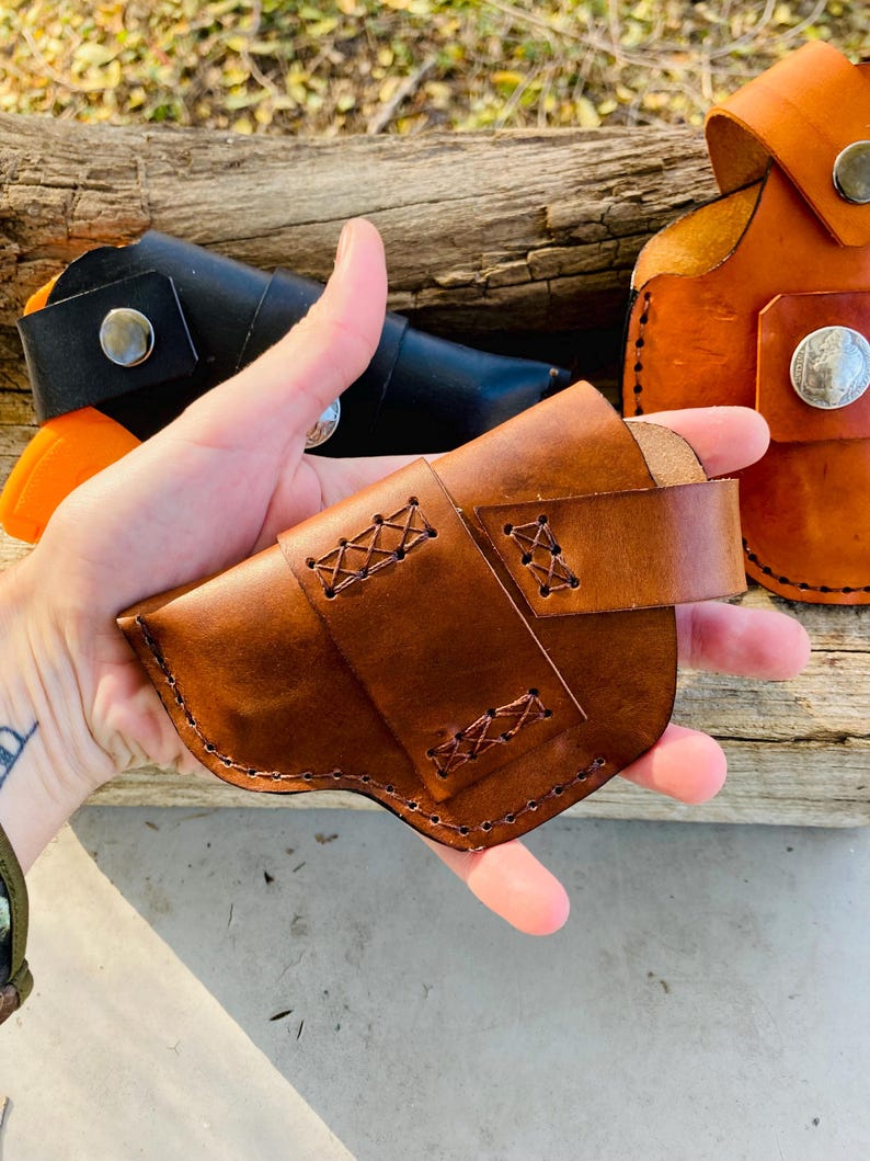 Op de afbeelding: Een bruin leren holster met decoratieve stiksels en een riem. Andere holsters in zwart en oranje zijn zichtbaar op de achtergrond. De holsters zijn gemaakt van leer en hebben metalen knoopsluitingen. De stiksels zijn zichtbaar.