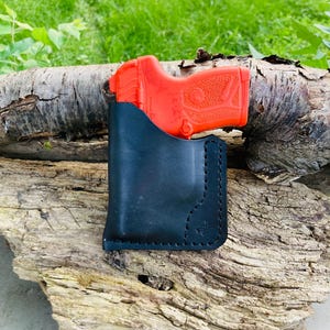May include: A black leather holster with a red handgun inside, resting on a piece of weathered wood. The holster is stitched and features a decorative emblem. The background is blurred green foliage.