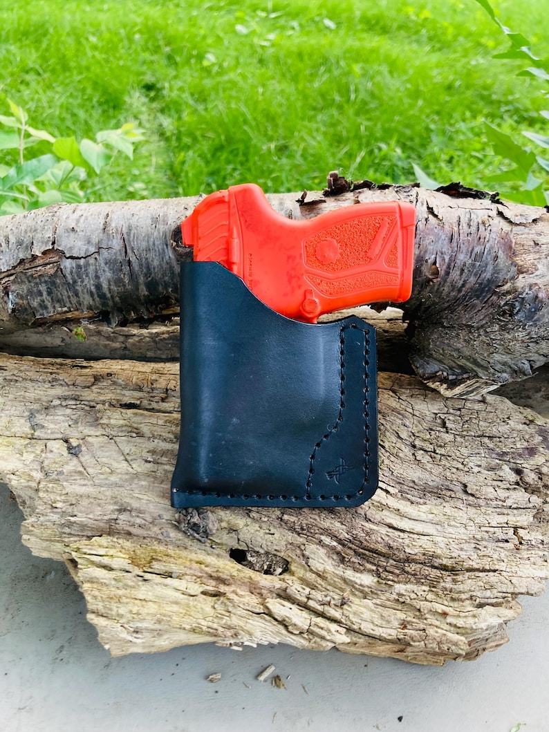 May include: A black leather holster with a red handgun inside, resting on a piece of weathered wood. The holster is stitched and features a decorative emblem. The background is blurred green foliage.