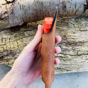 K&ouml;nnte beinhalten: Eine braune Lederscheide mit einem leuchtend orangenen Gegenstand im Inneren. Die Scheide hat eine spitze Spitze und wird in einer Hand gehalten.