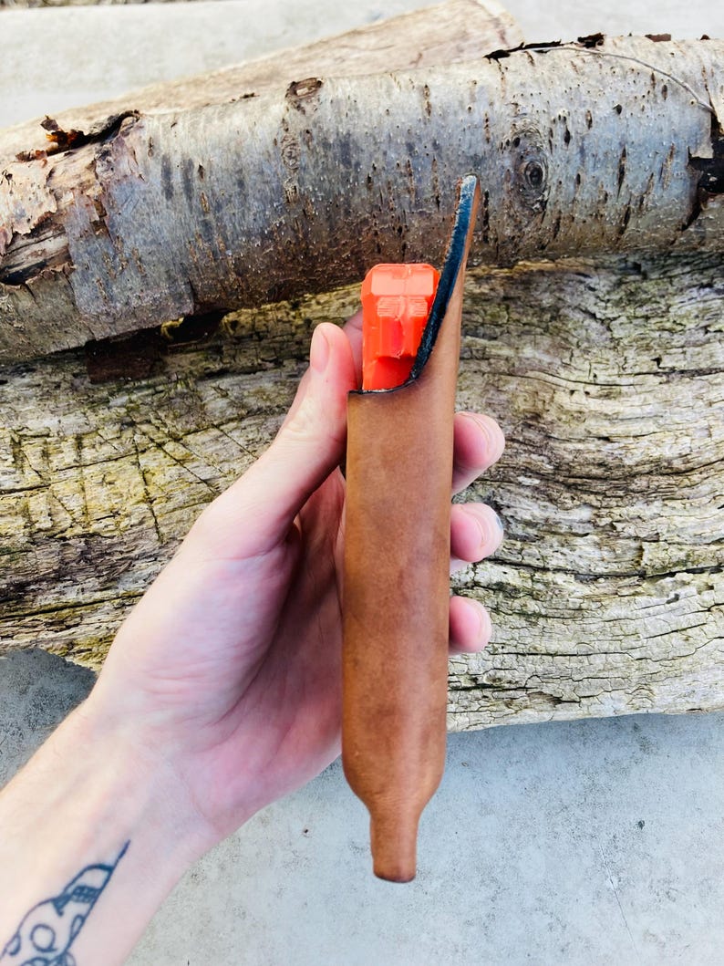 K&ouml;nnte beinhalten: Eine braune Lederscheide mit einem leuchtend orangenen Gegenstand im Inneren. Die Scheide hat eine spitze Spitze und wird in einer Hand gehalten.