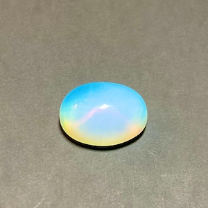 Puede incluir: Una piedra preciosa de ópalo ovalada y pulida. La piedra muestra un degradado de colores, pasando del azul en la parte superior al amarillo en la inferior, con un brillo nacarado. La gema está sobre un fondo gris.