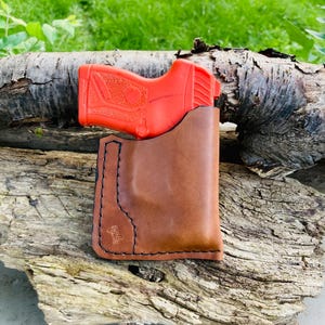 May include: A brown leather holster with black stitching contains a bright orange handgun. The holster is placed on a weathered piece of wood, with a blurred green background of grass and foliage.