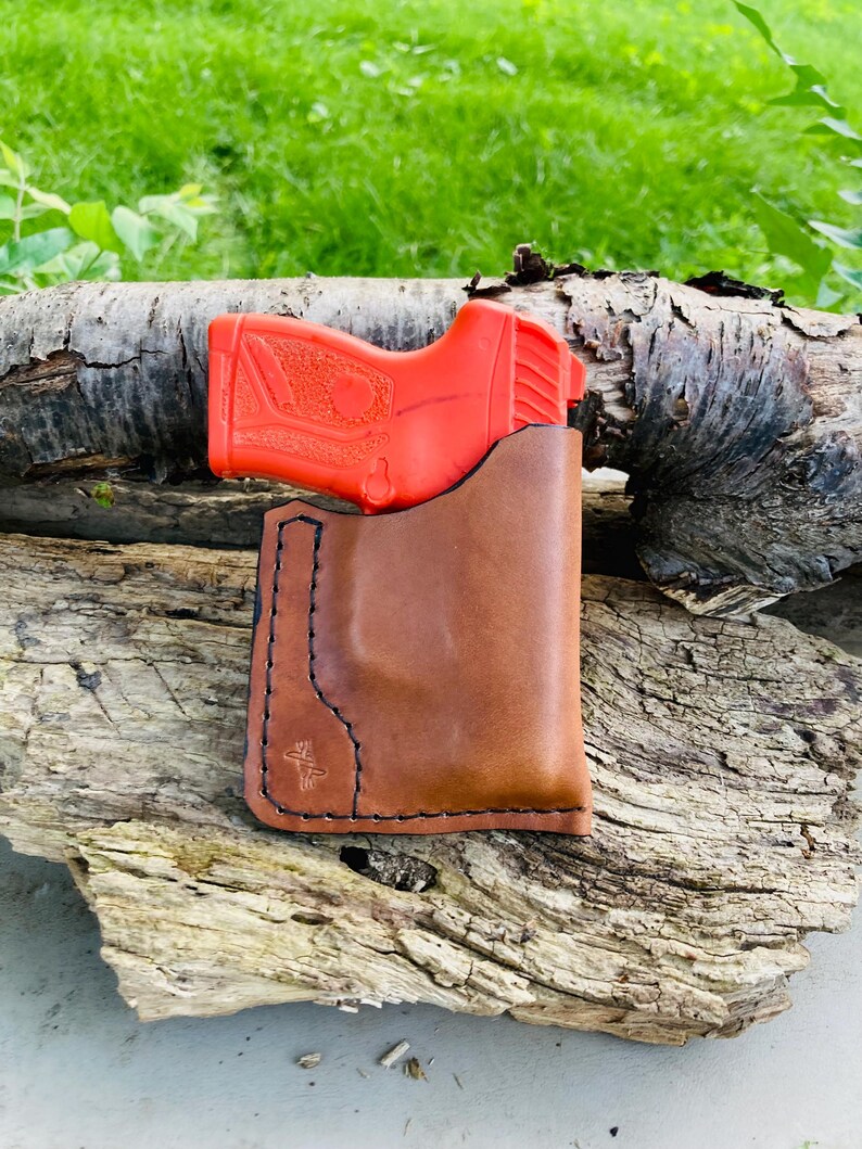 May include: A brown leather holster with black stitching contains a bright orange handgun. The holster is placed on a weathered piece of wood, with a blurred green background of grass and foliage.