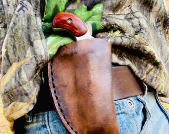 Könnte beinhalten: Ein kleines Jagdmesser mit Holzgriff und Lederscheide. Das Messer steckt in einer braunen Lederscheide, die an einem Gürtel befestigt ist. Die Person trägt eine Tarnjacke und eine blaue Jeans.