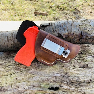 May include: A brown leather holster with a white stitch detail and a silver metal clip. A bright orange handgun is partially visible in the holster.
