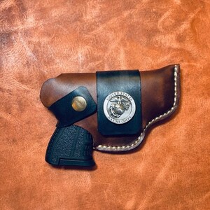 Op de afbeelding: Een bruine leren holster met een zwart pistool erin. De holster heeft een embleem van het United States Marine Corps op de voorkant.