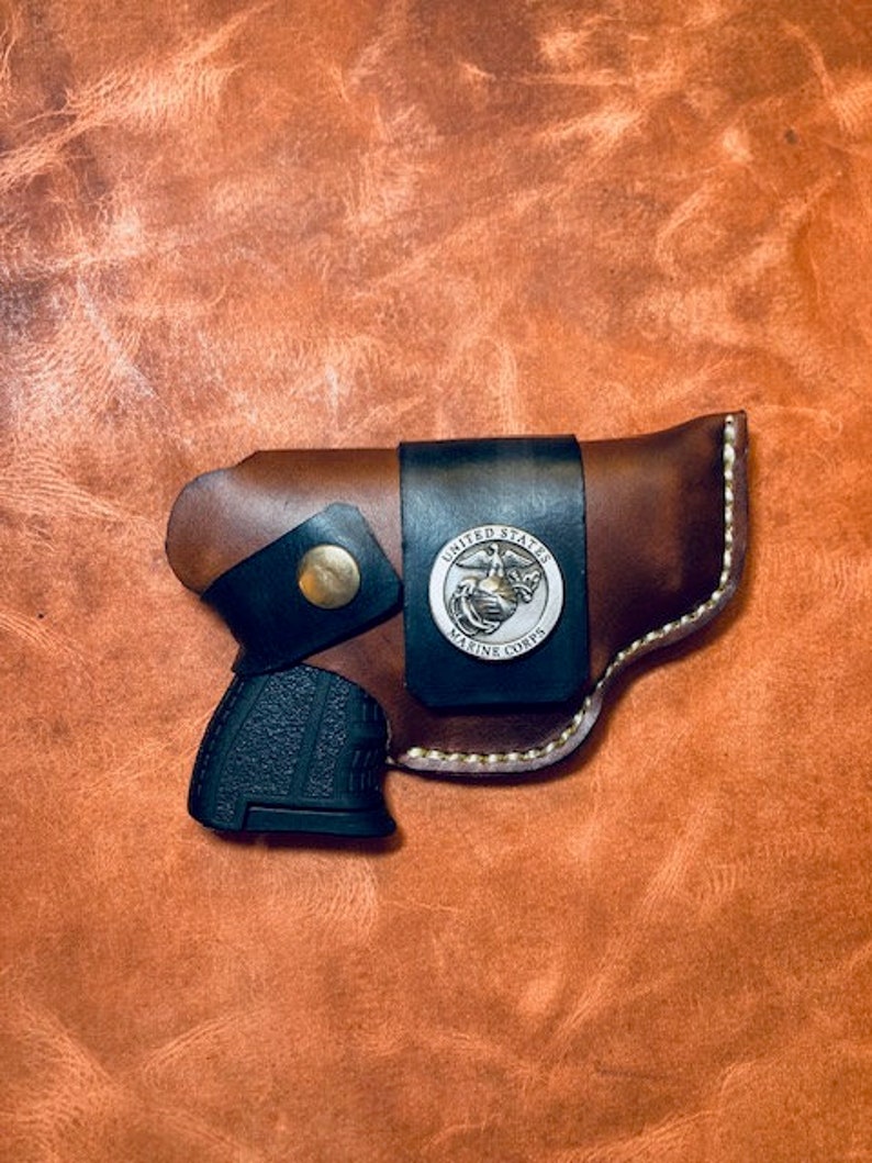 Op de afbeelding: Een bruine leren holster met een zwart pistool erin. De holster heeft een embleem van het United States Marine Corps op de voorkant.