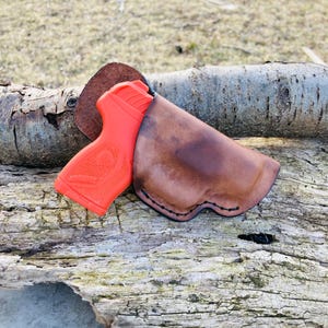 K&ouml;nnte beinhalten: Ein braunes Lederholster mit einer leuchtend orangefarbenen Pistole darin. Das Holster ist mit schwarzem Faden vern&auml;ht und hat einen leicht abgenutzten Look. Das Holster liegt auf einem St&uuml;ck verwittertem Holz.