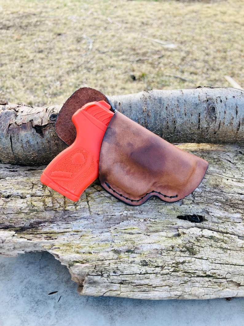 K&ouml;nnte beinhalten: Ein braunes Lederholster mit einer leuchtend orangefarbenen Pistole darin. Das Holster ist mit schwarzem Faden vern&auml;ht und hat einen leicht abgenutzten Look. Das Holster liegt auf einem St&uuml;ck verwittertem Holz.