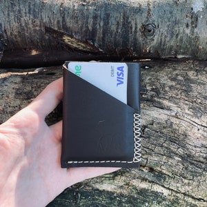 Puede incluir: Una cartera de cuero marr&oacute;n con un borde cosido y una ranura para tarjeta. La cartera contiene una tarjeta de d&eacute;bito blanca y verde con el logotipo de VISA y la palabra "DEBIT".