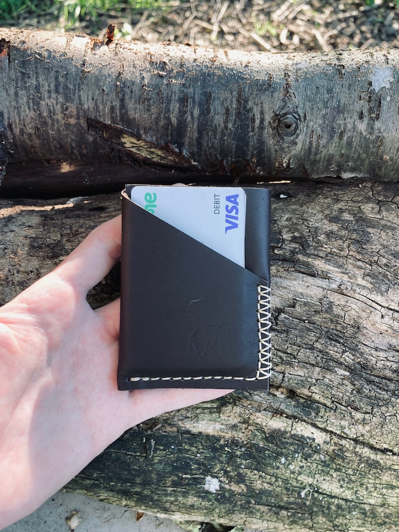Puede incluir: Una cartera de cuero marr&oacute;n con un borde cosido y una ranura para tarjeta. La cartera contiene una tarjeta de d&eacute;bito blanca y verde con el logotipo de VISA y la palabra "DEBIT".