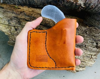 Könnte beinhalten: Eine hellbraune Lederscheide mit einer klaren, gebogenen Klinge, die teilweise eingesetzt ist. Die Scheide hat einen genähten Rand und eine geprägte amerikanische Flagge. Der Hintergrund ist ein Stück verwittertes Holz.