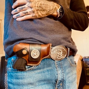 Op de afbeelding: Een bruine leren holster met een zwarte band en een zilveren gesp. De holster is bevestigd aan een blauwe jeans met een zilveren gesp die "Don't Tread On Me" zegt en een slangontwerp heeft.