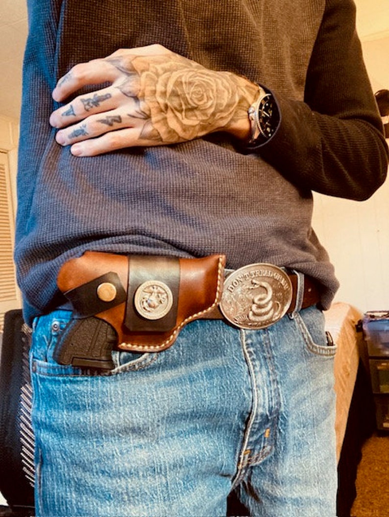 Op de afbeelding: Een bruine leren holster met een zwarte band en een zilveren gesp. De holster is bevestigd aan een blauwe jeans met een zilveren gesp die "Don't Tread On Me" zegt en een slangontwerp heeft.